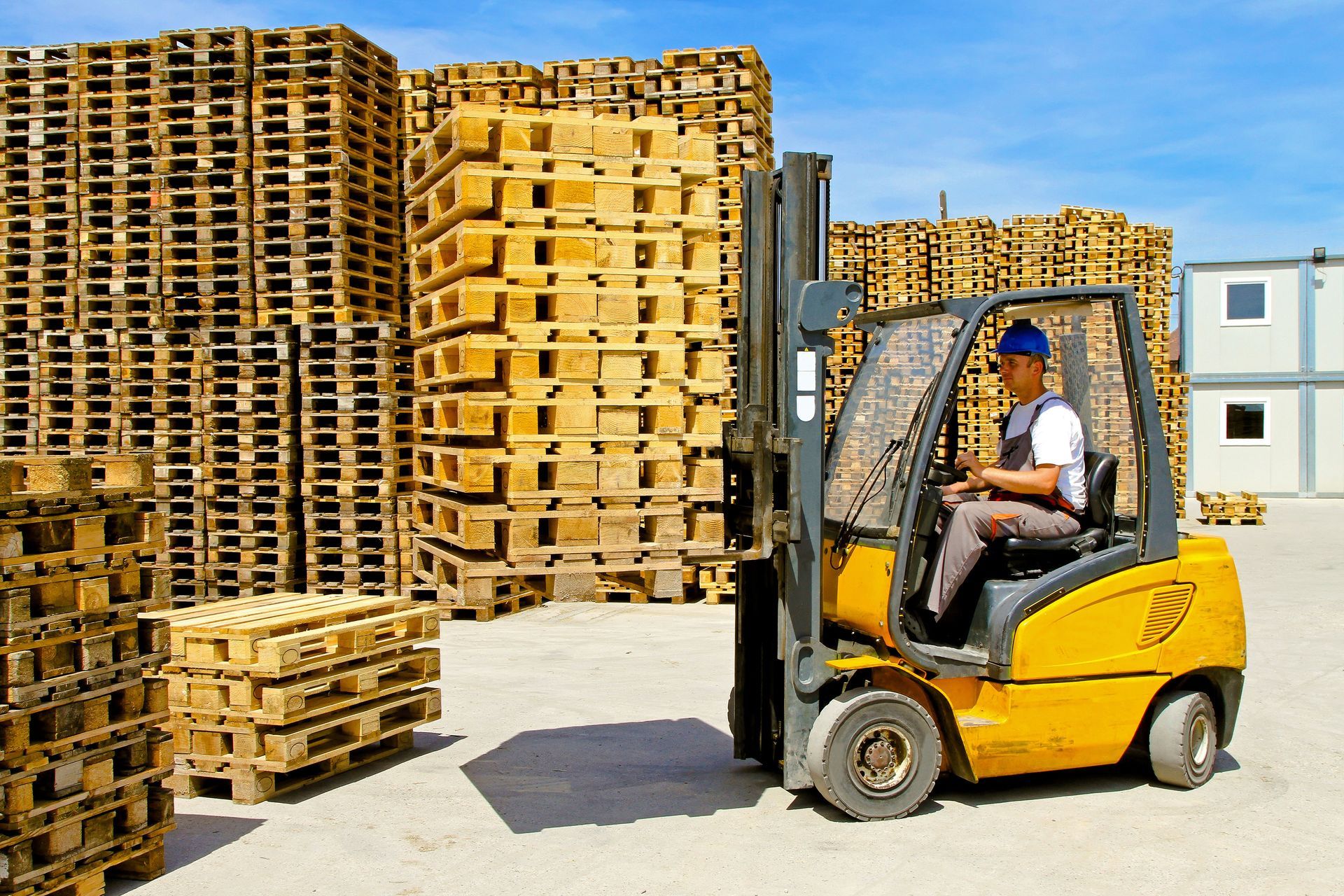 Forklift operator moving wooden pallets in a storage yard under a blue sky.