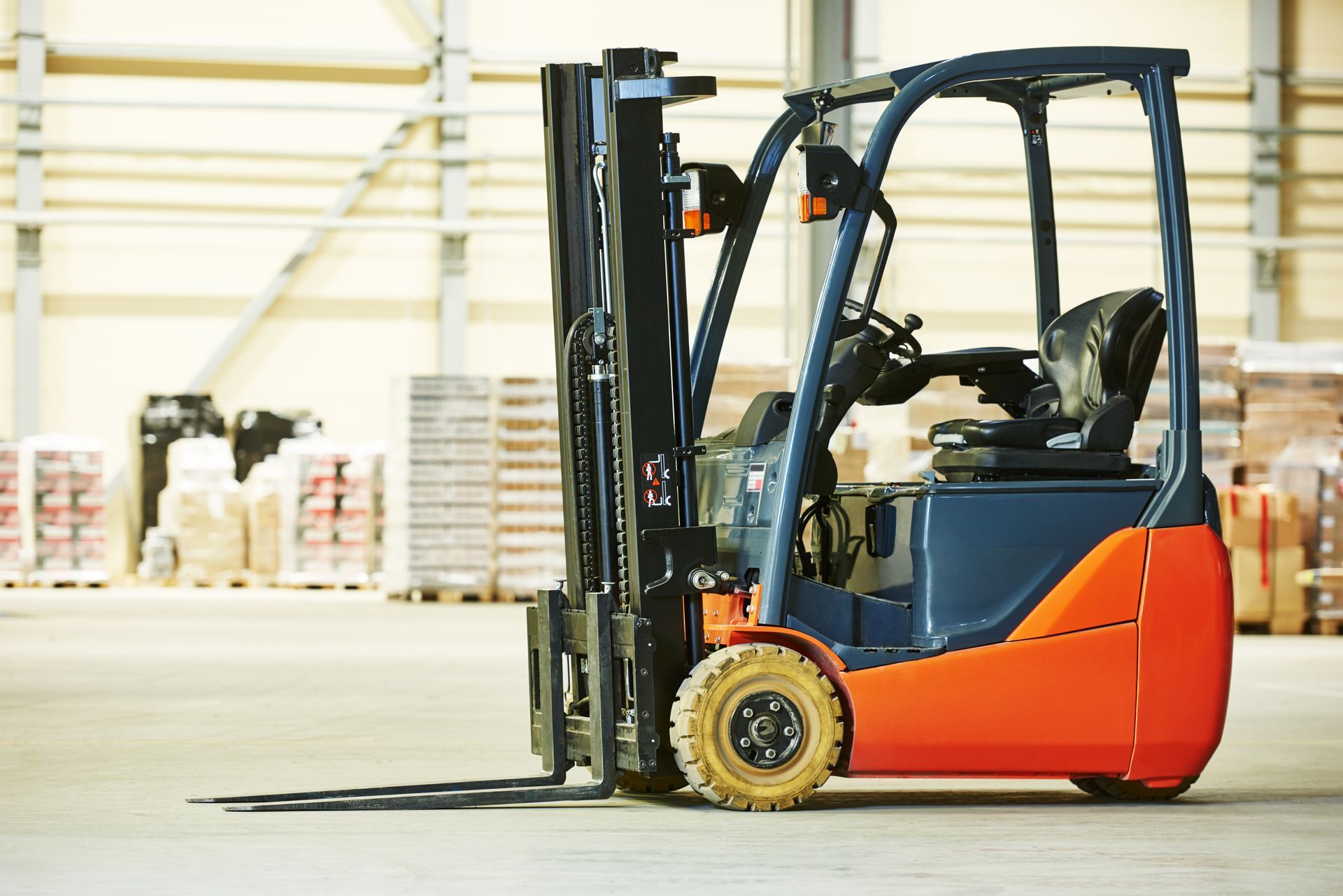 Forklift in warehouse; blue and orange, ready for use, pallets in background.