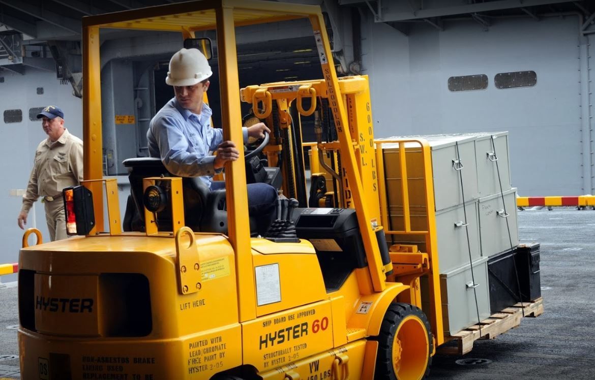 Forklift operator moving wooden pallets in a storage yard under a blue sky.