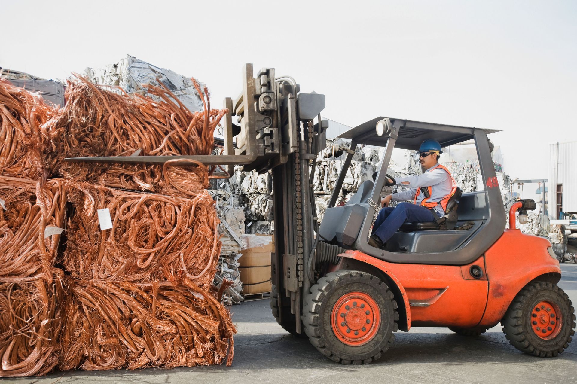 Forklift operator moving a large stack of copper scrap. Orange forklift, blue hard hat, outdoor setting.