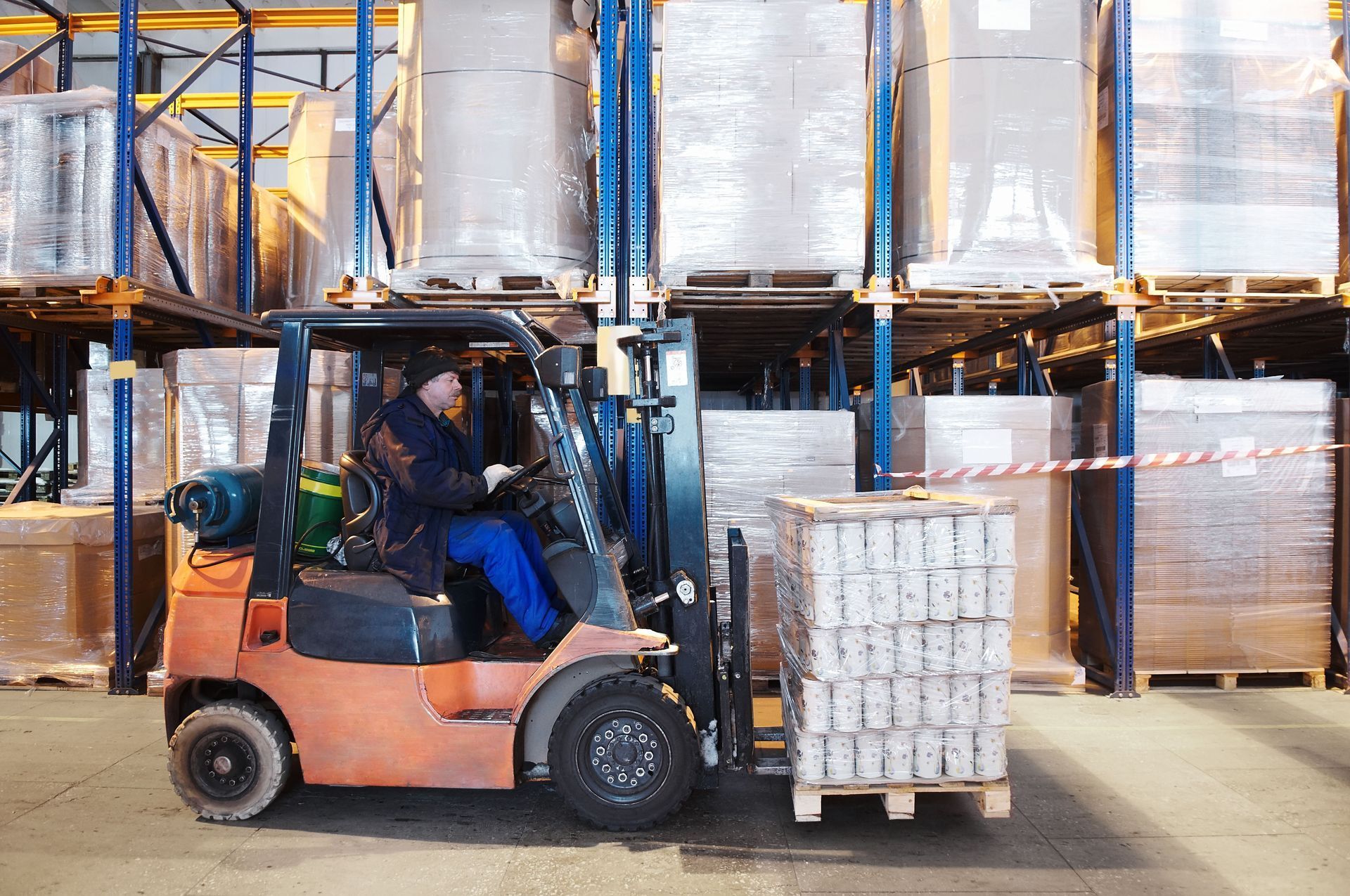 Forklift operator in a warehouse moves a pallet of items. Shelves are stacked with boxes in the background.