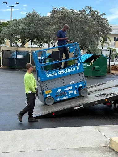 Two workers loading a blue Genie lift onto a flatbed truck; one is operating the lift, the other is walking beside it.
