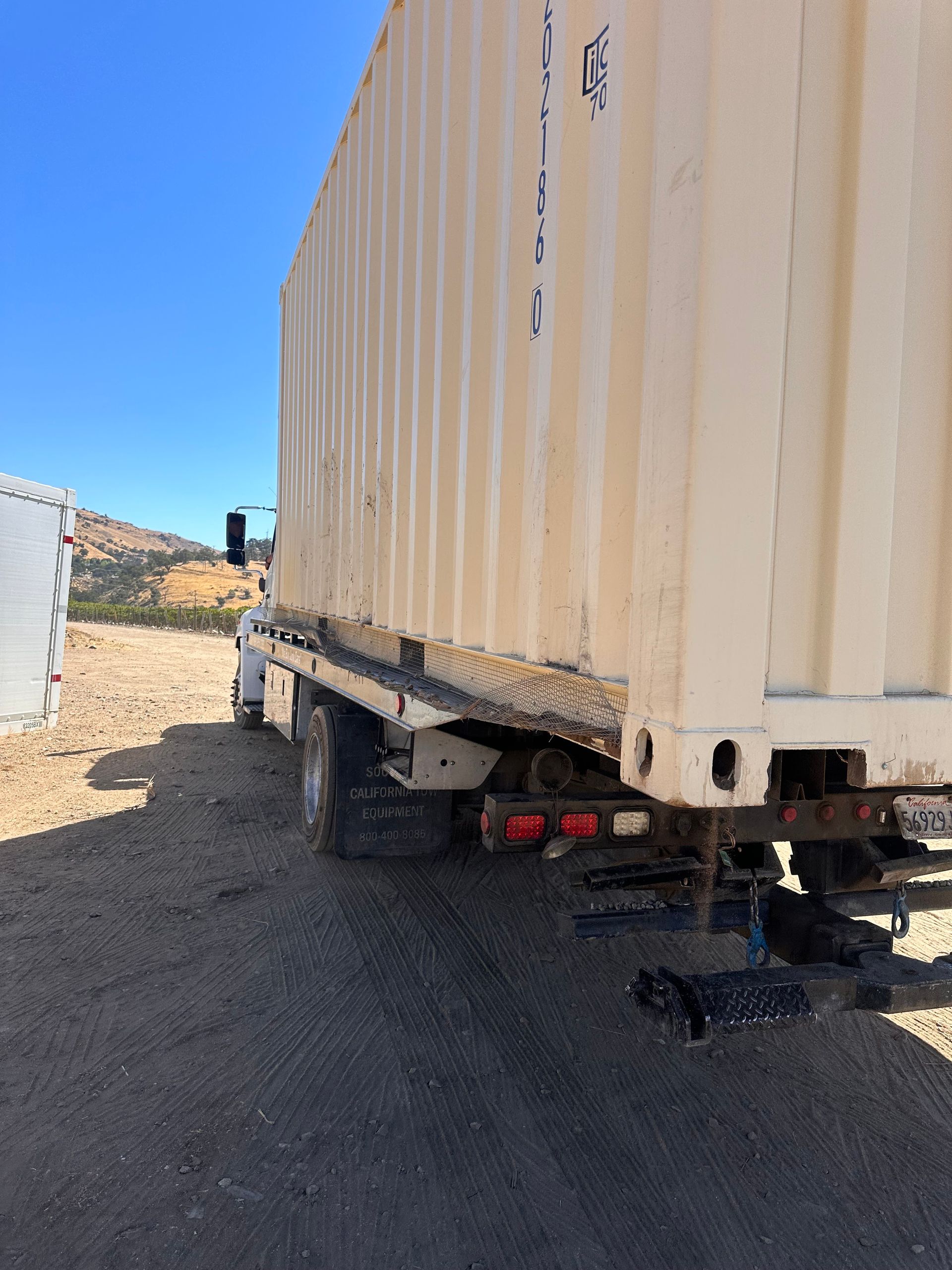 Tan shipping container on a flatbed truck in a dirt lot on a sunny day.