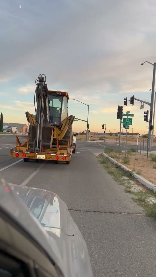 A backhoe on a trailer being driven on a road at sunset.