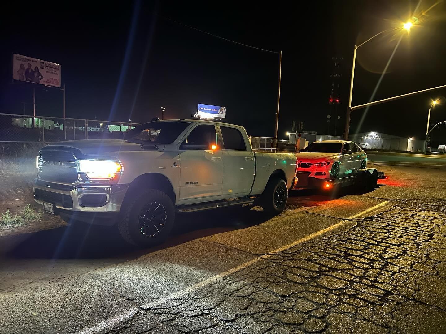 White pickup truck towing a silver car on a cracked road at night. Headlights and streetlights illuminate the scene.