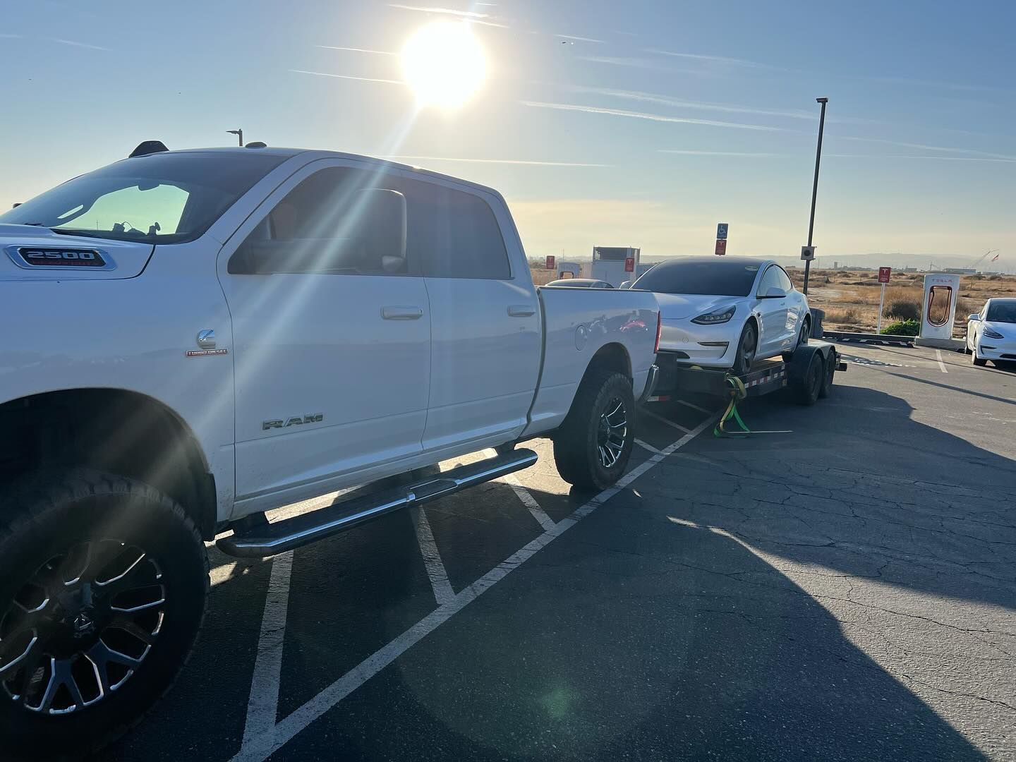 White truck towing a white Tesla on a trailer in a charging station parking lot under a bright sun.
