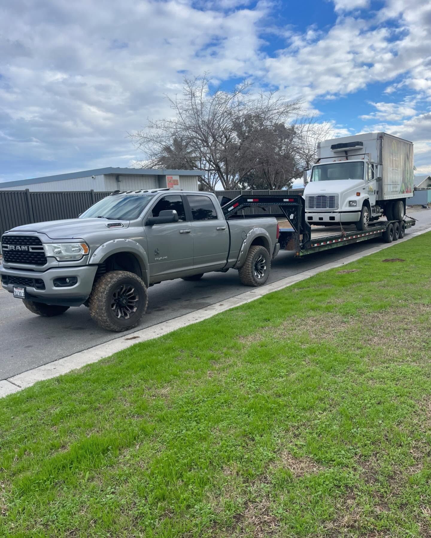 Gray truck towing a white box truck on a trailer along a road with green grass.