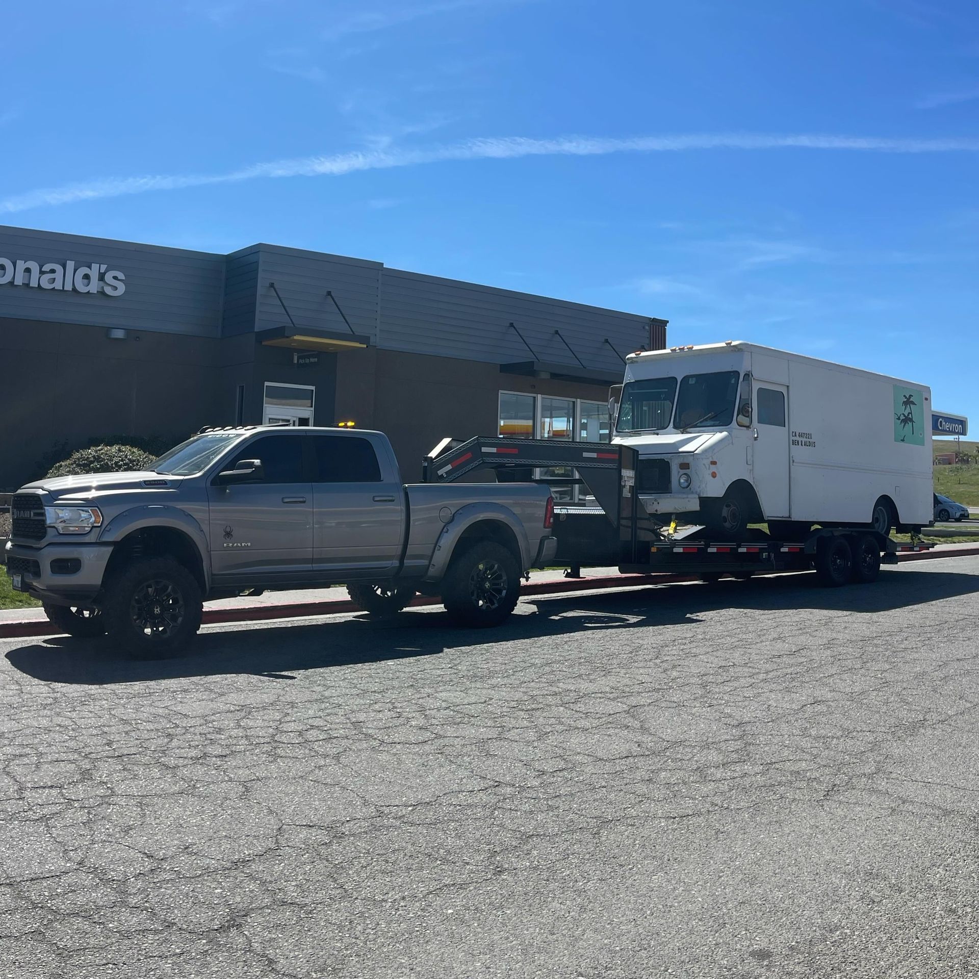 Grey pickup truck towing a white box truck on a flatbed trailer in front of a McDonald's restaurant.