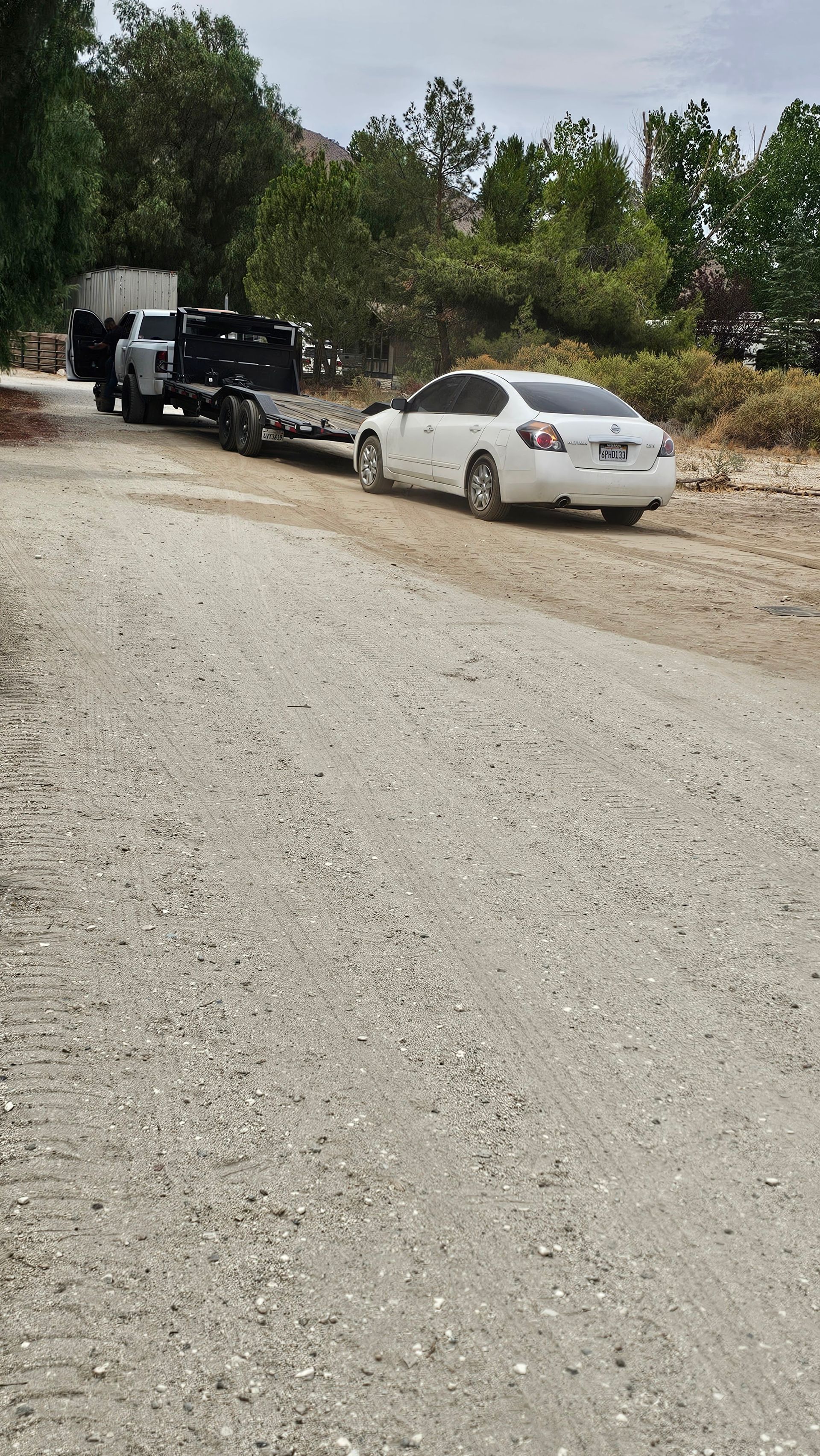 White car towing a trailer carrying a black jeep and a white truck on a gravel road, trees in the background.
