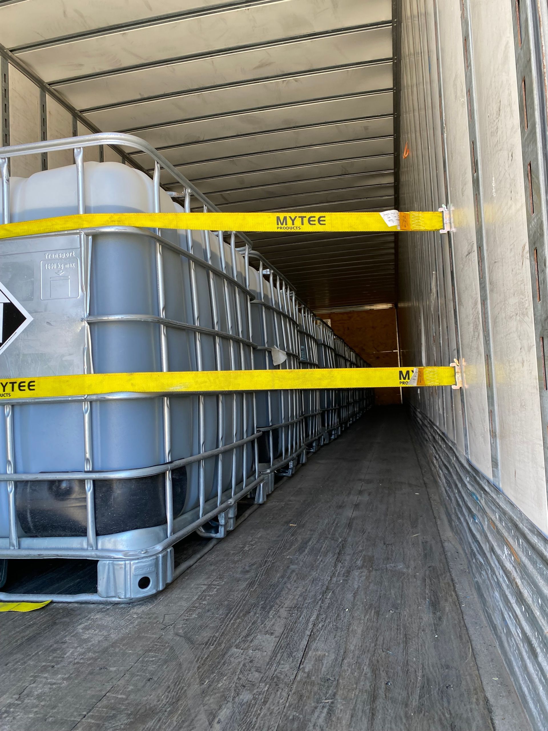 Inside a truck trailer, rows of gray liquid totes are secured with yellow bars.