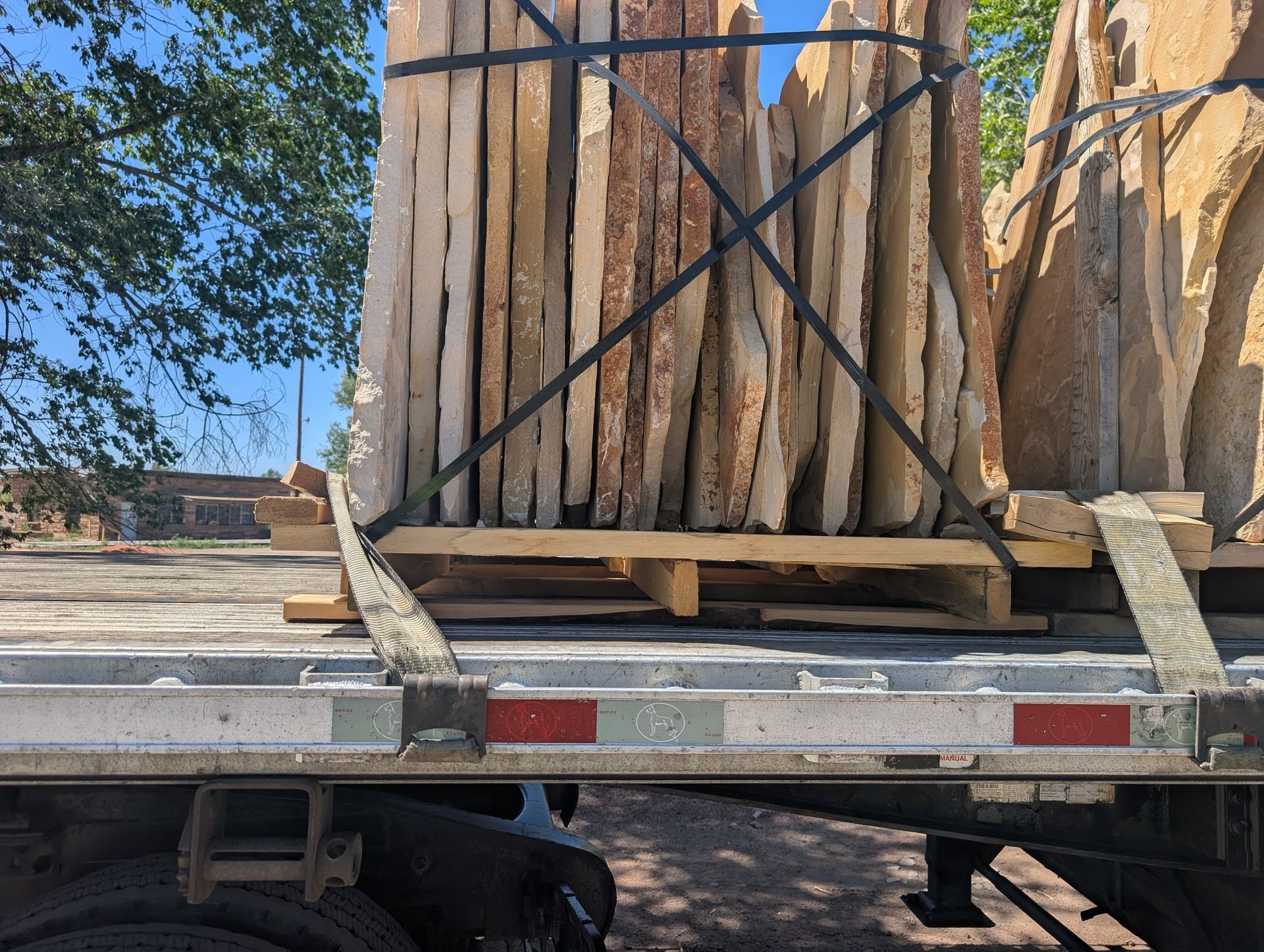 Pallet of tan flagstone secured on a flatbed trailer. Straps and the sky are visible.
