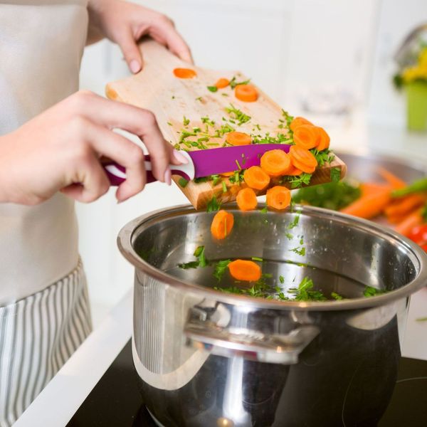 Person adding chopped carrots and herbs from cutting board into a stainless steel pot on a stove.