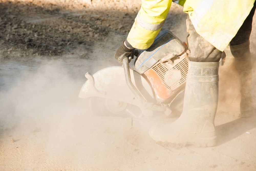 Person in safety gear using a concrete saw, creating a cloud of dust.