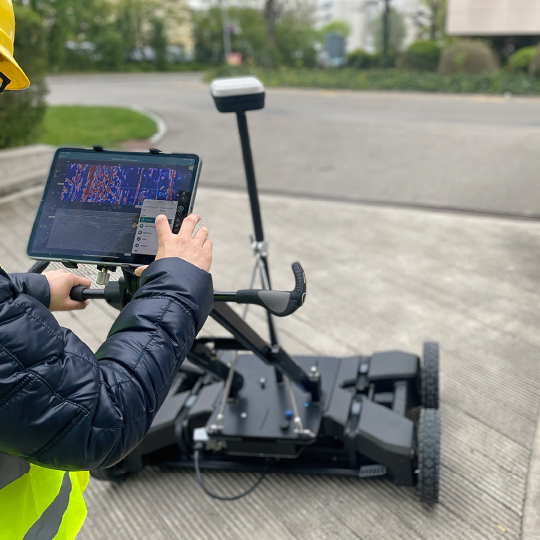 Person operating a remote-controlled mower on a paved path outdoors, wearing a high-vis vest.