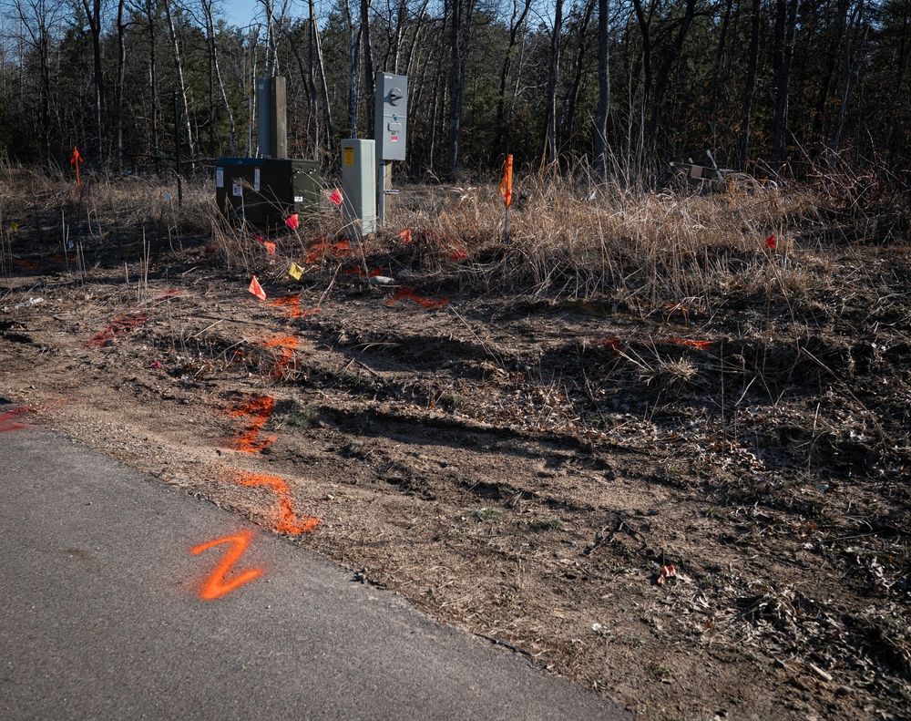 Orange survey markings on a dirt roadside near utility boxes and a wooded area