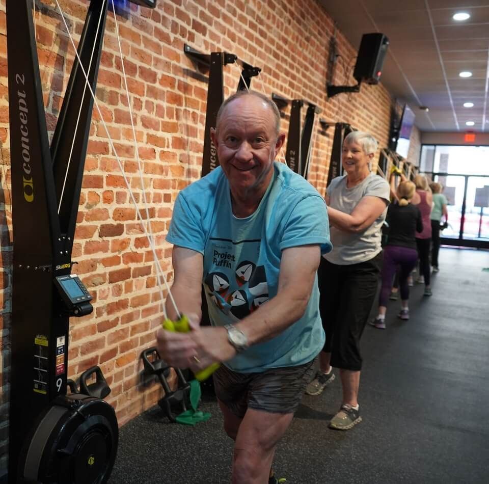 A person in a gym in New Orleans sits on a bench talking to another person standing next to a rowing machine.