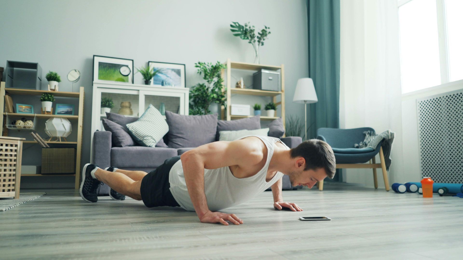 Person doing a push-up on a yoga mat in a bright living room with plants and a chair nearby