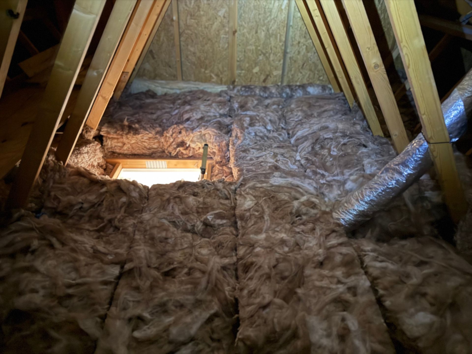 Attic space with brown insulation between wooden beams and a small open window.