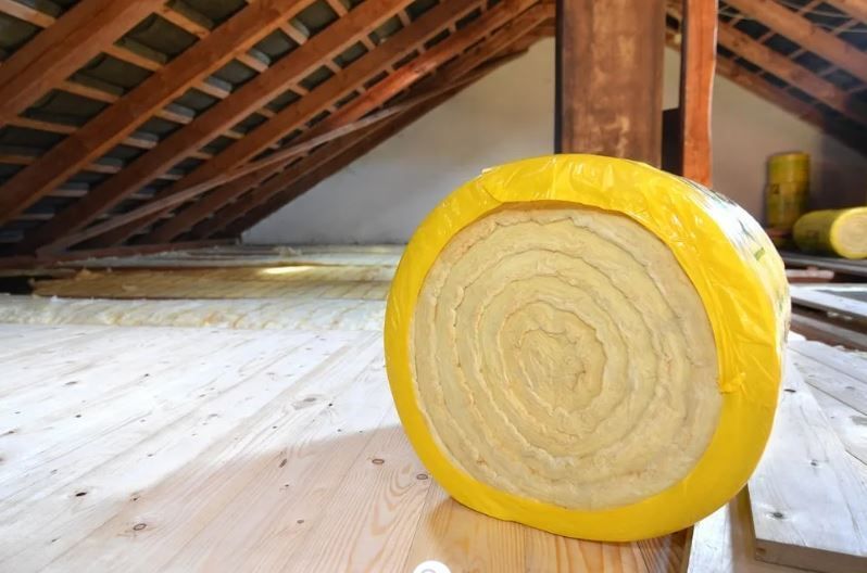 Roll of yellow insulation in an attic, wooden beams above, on a wooden floor.
