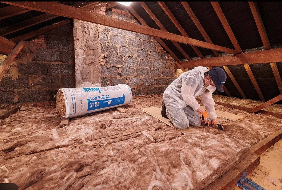 Person in protective suit cuts insulation in an Loft, with a roll of insulation nearby.