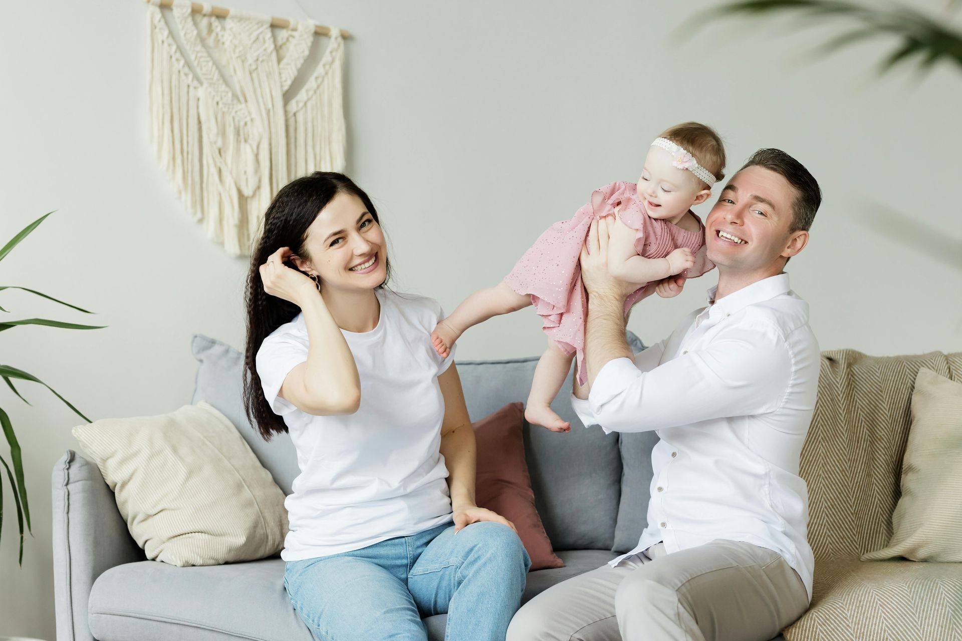 Family on a couch. Father holding up a baby; mother smiles. Bright living room setting.