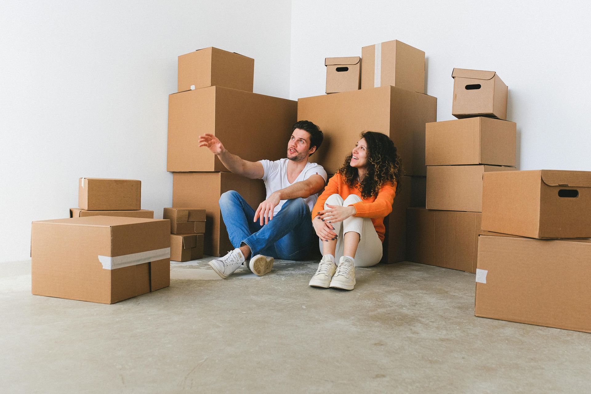 Couple sitting amongst moving boxes, man pointing, woman looking up. Interior, light-colored walls.