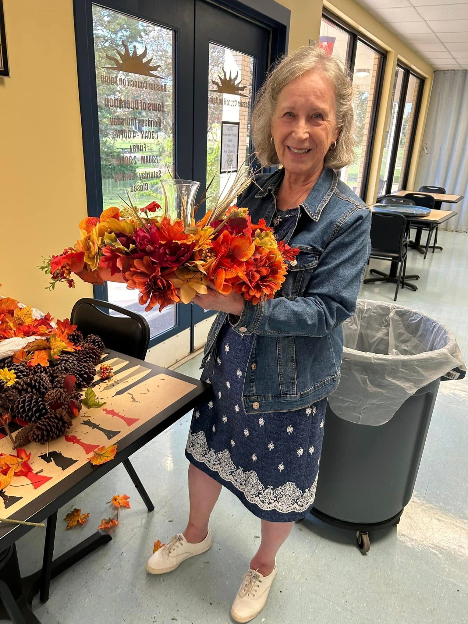 Woman holding autumn floral arrangement; stands near tables, trash can, and window; smiling.