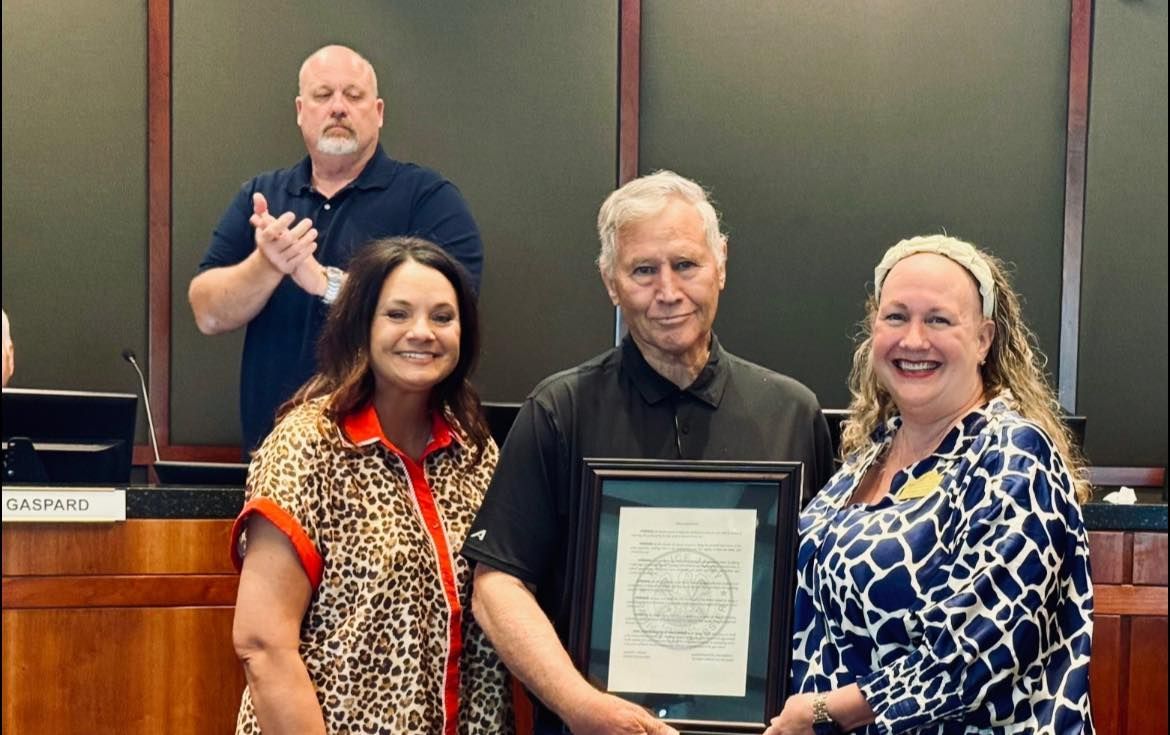 Three people hold a framed document; a man in black is in the middle, two women flank him and a man claps in the background.