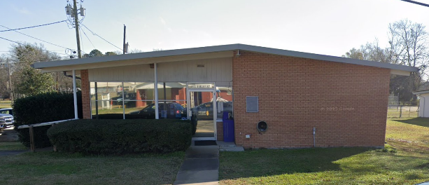 A brick building with glass windows, a covered walkway, and a row of bushes in front.