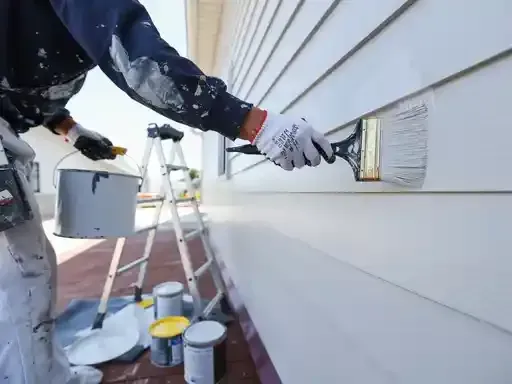 Painter applying white paint to a house exterior. White paint bucket, ladder, and paint cans present.