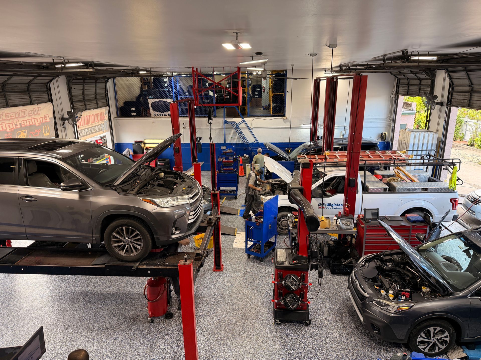 Cars on lifts in a busy auto repair shop, various tools and equipment visible, open garage doors.