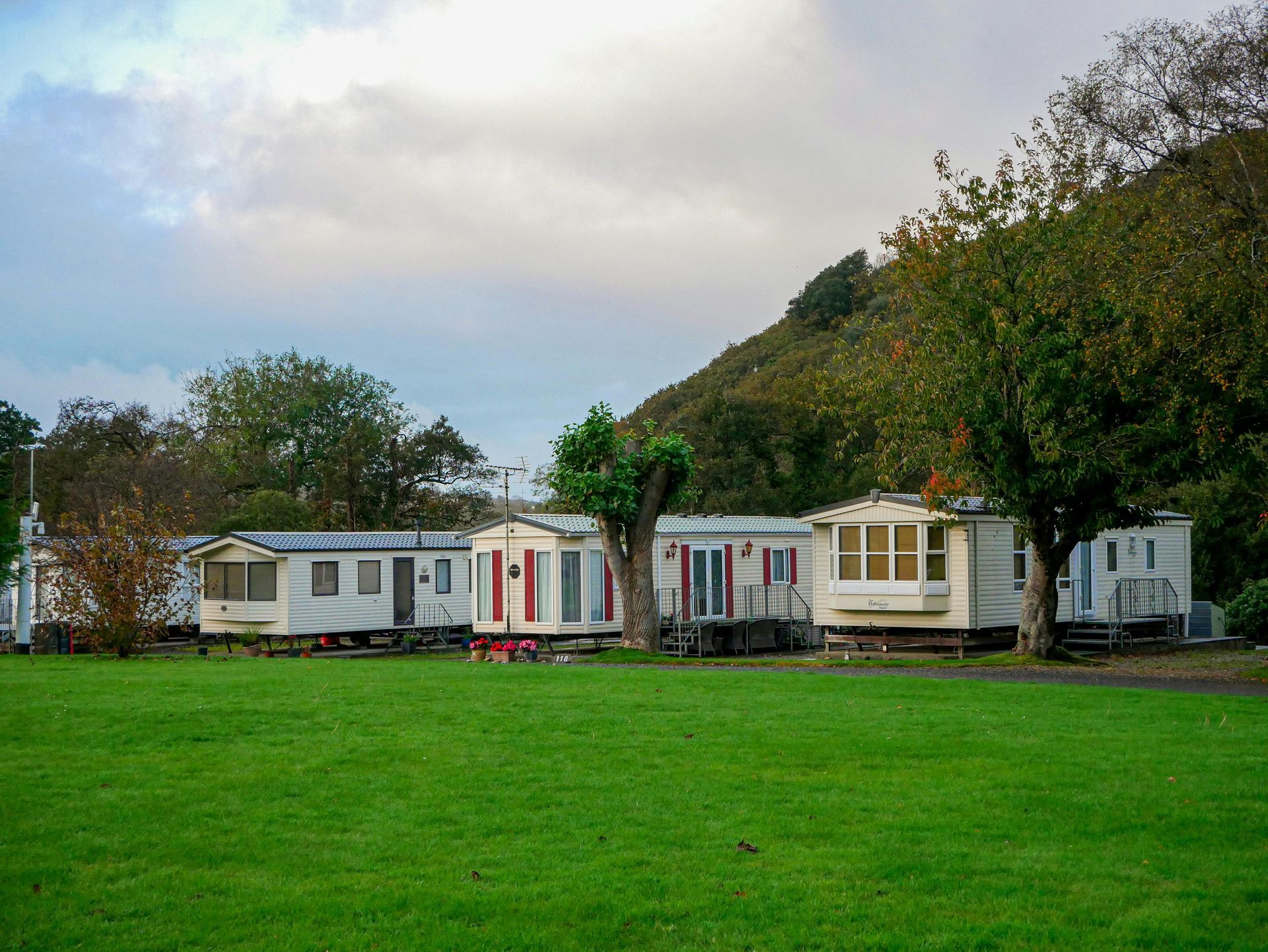 Mobile homes in a green field with trees and a hill under a cloudy sky.