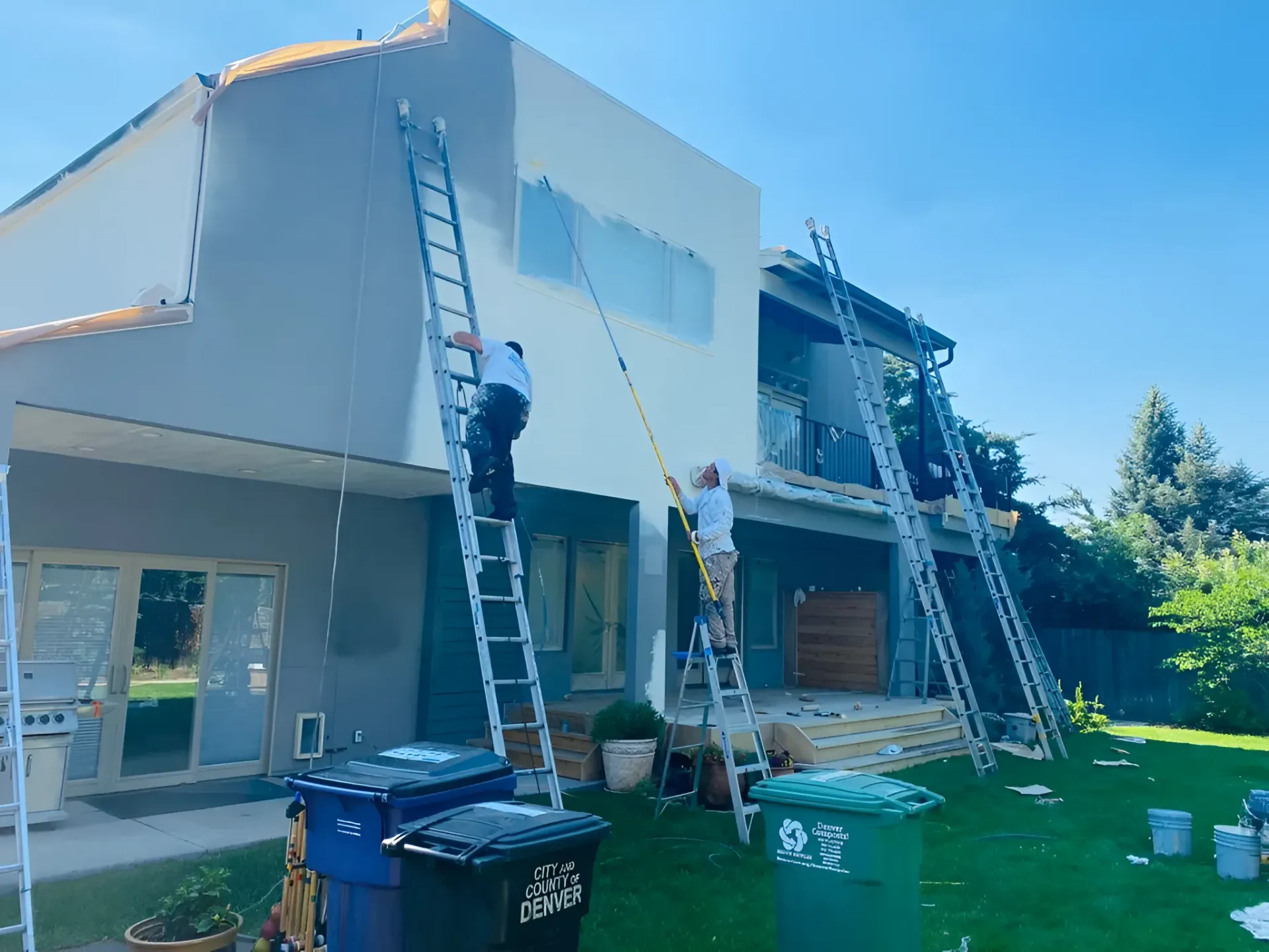 Men on ladders painting a two-story house exterior; gray and white walls, blue skies, green lawn.