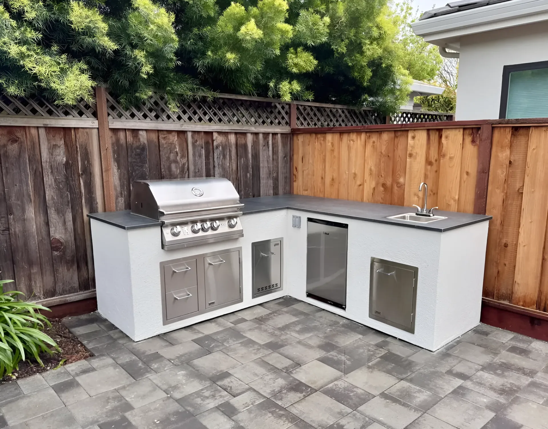 Outdoor L-shaped kitchen with grill, sink, and refrigerator on a gray stone patio beside a wooden fence.