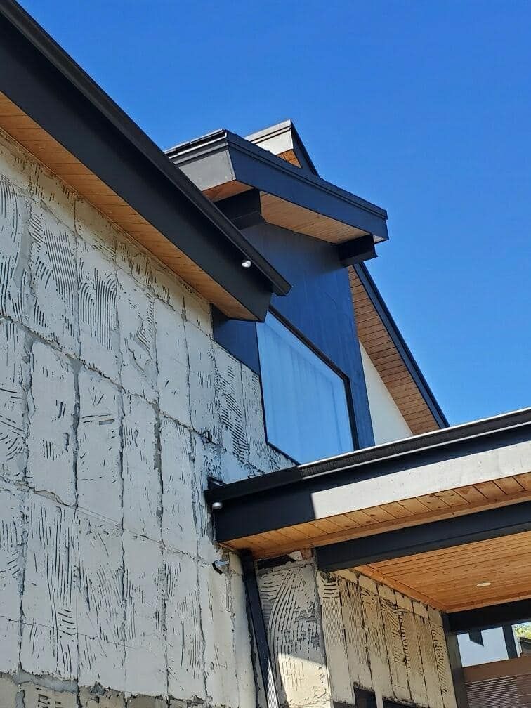 Exterior of a house under construction. Light-colored concrete wall, dark roof and trim, blue sky.
