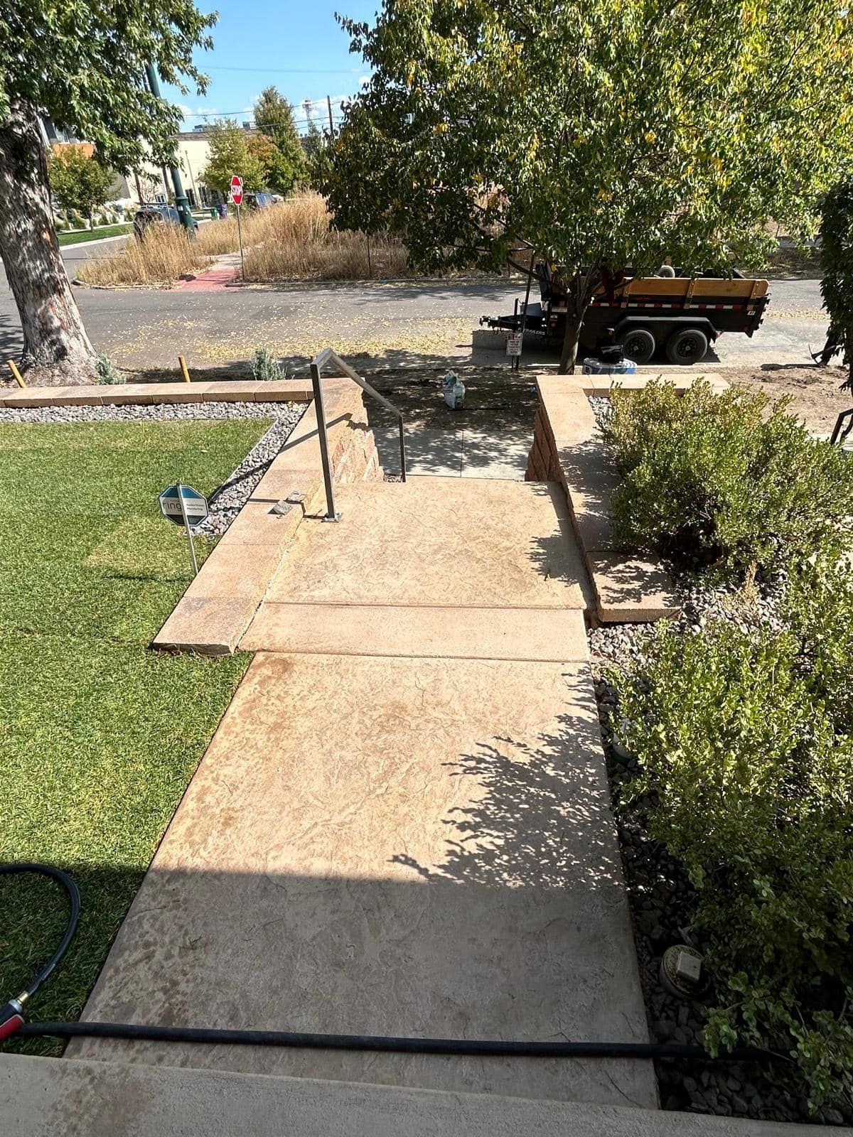 Concrete walkway with grass, bushes, and a tree. A trailer is visible in the background.