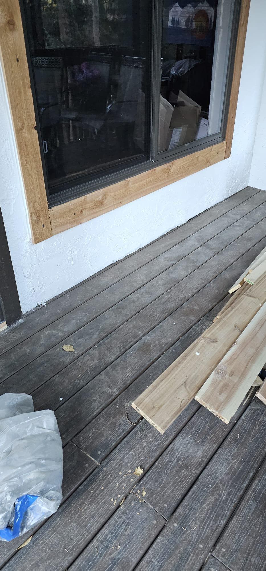 Exterior view of a deck with weathered boards. A window frame is visible. Unfinished wooden planks are nearby.