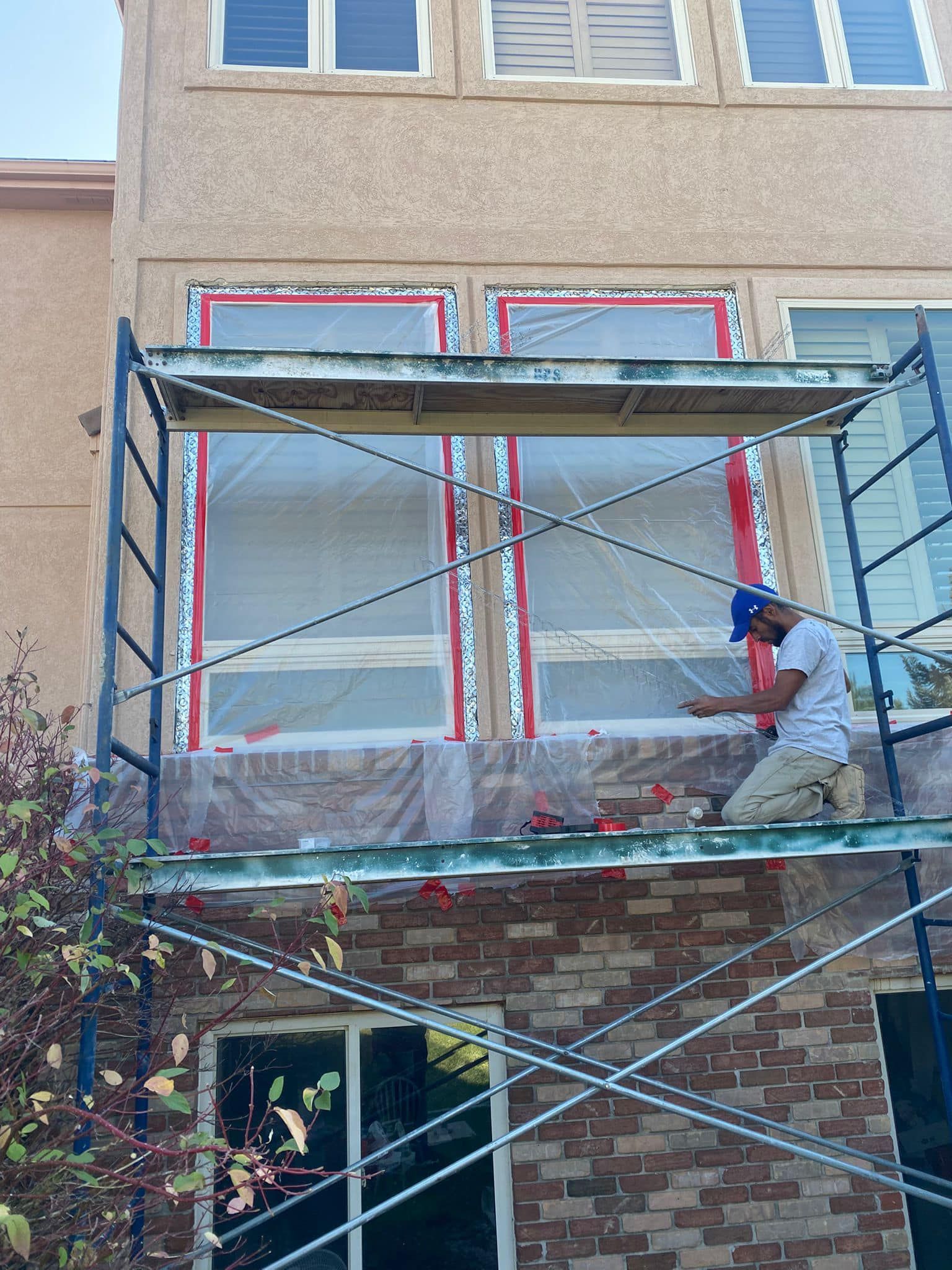 Man on scaffolding, bricklaying near windows taped with red masking tape and plastic wrap. Building exterior.