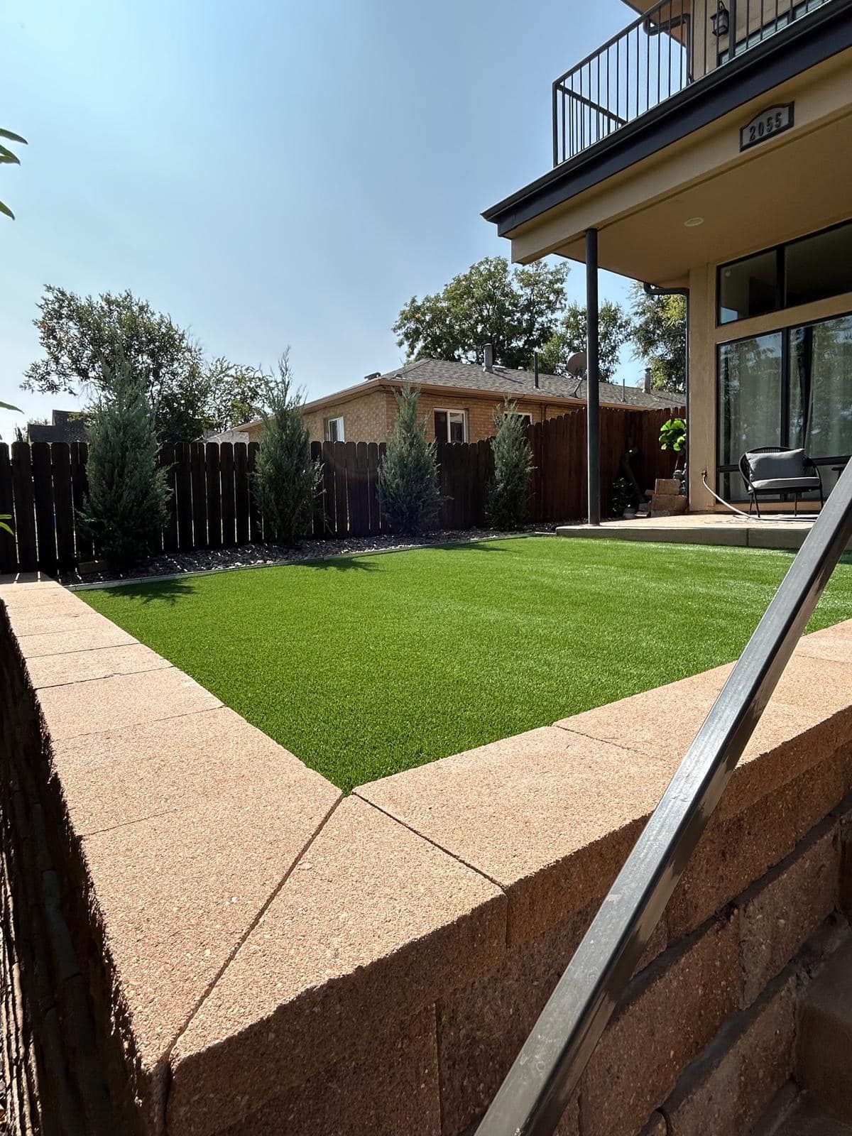 Backyard with a green lawn, retaining wall, fence, and a building with a balcony.