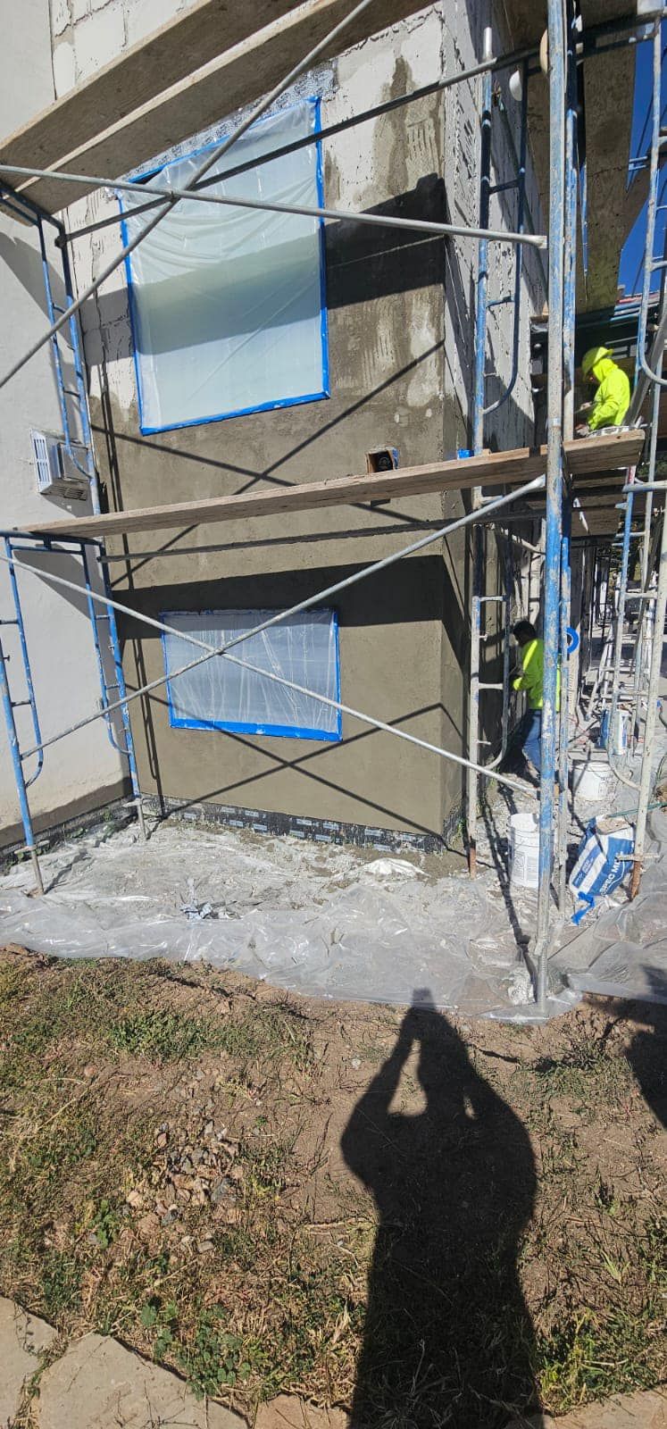 Construction workers applying stucco to a building exterior. Windows are taped off. Scaffolding is in place.