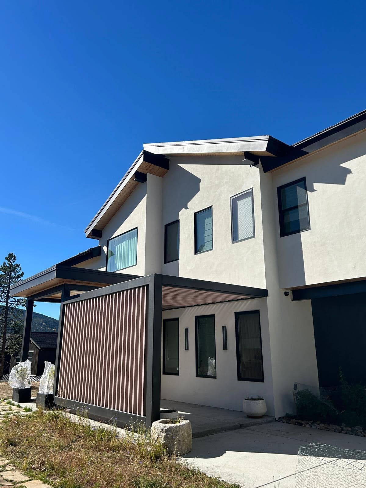 Two-story modern house with white stucco walls, black trim, and a pergola with brown slats under a bright blue sky.