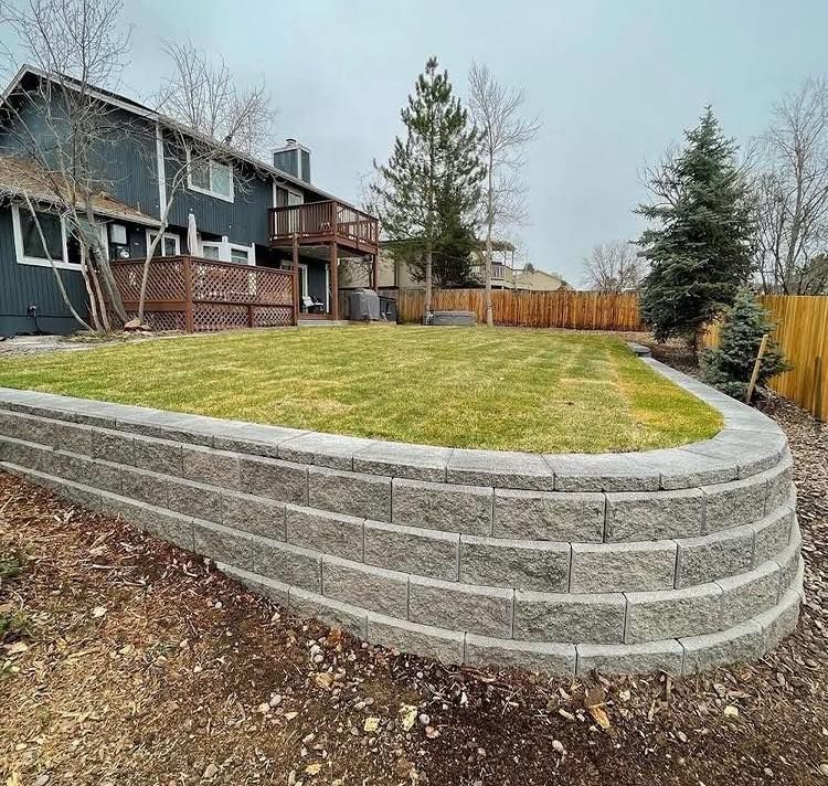 Gray retaining wall curves around a grassy yard in front of a blue house and wooden fence.