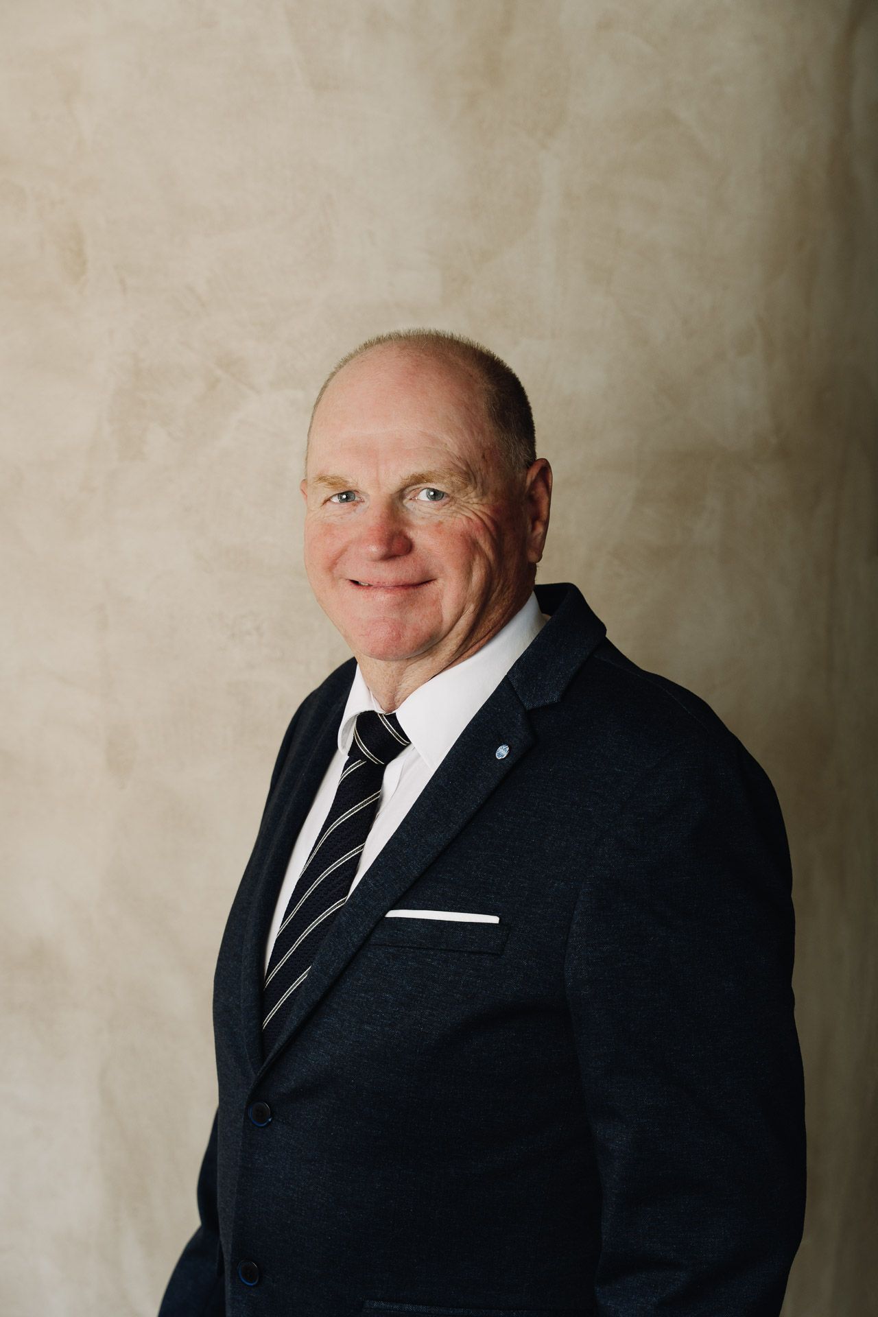 Man in a dark blazer, white shirt, and striped tie smiles. Pale background.