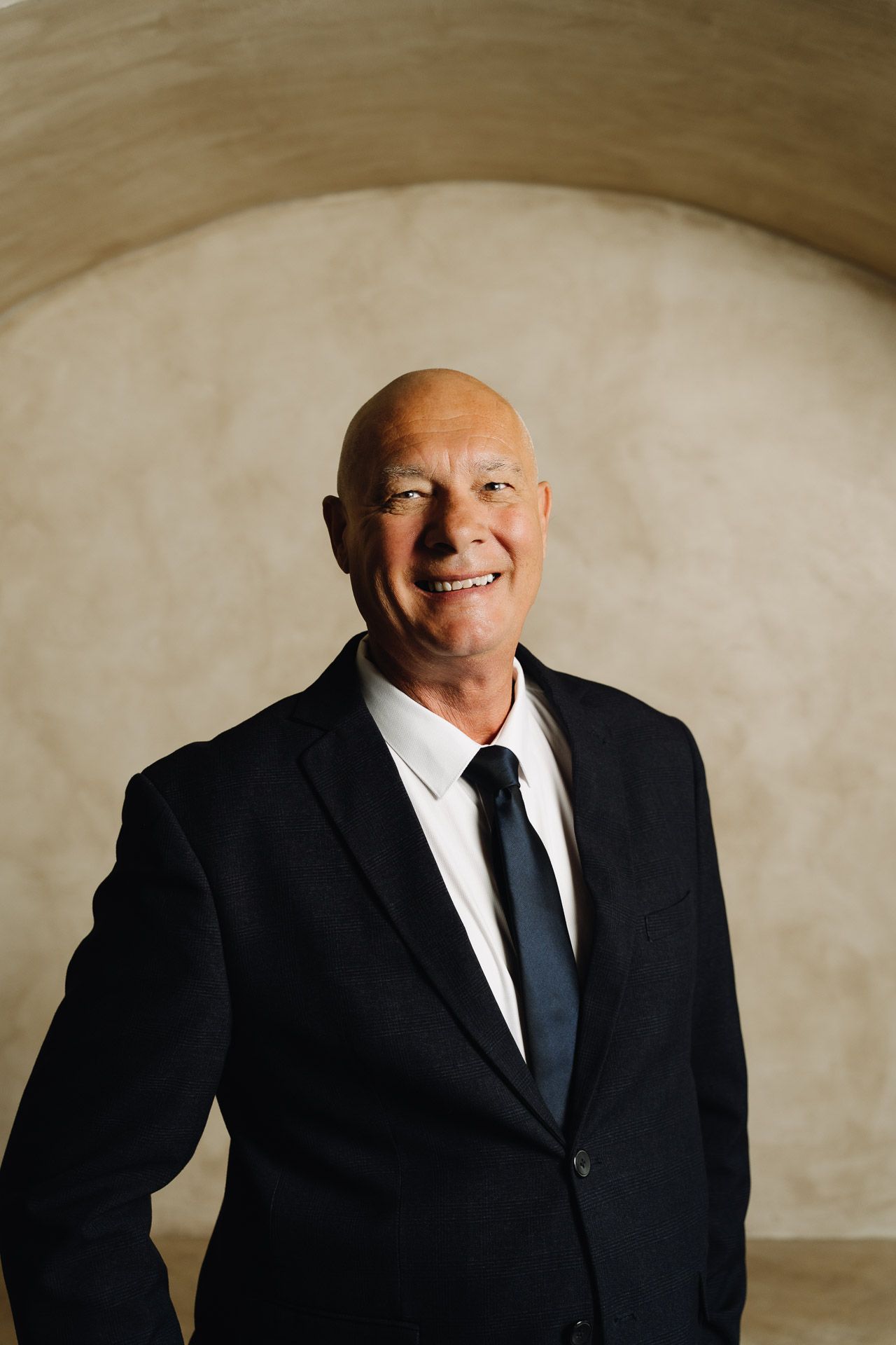 Bald man in a black suit smiles, standing against a beige arched wall.