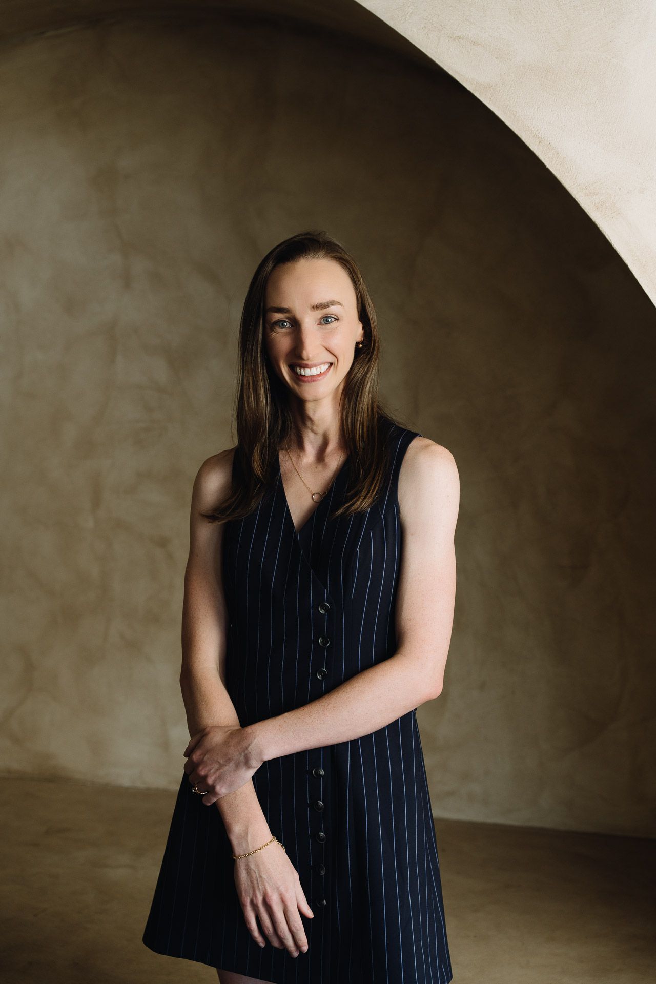 Woman in dark sleeveless dress smiles, stands near a tan arched wall.