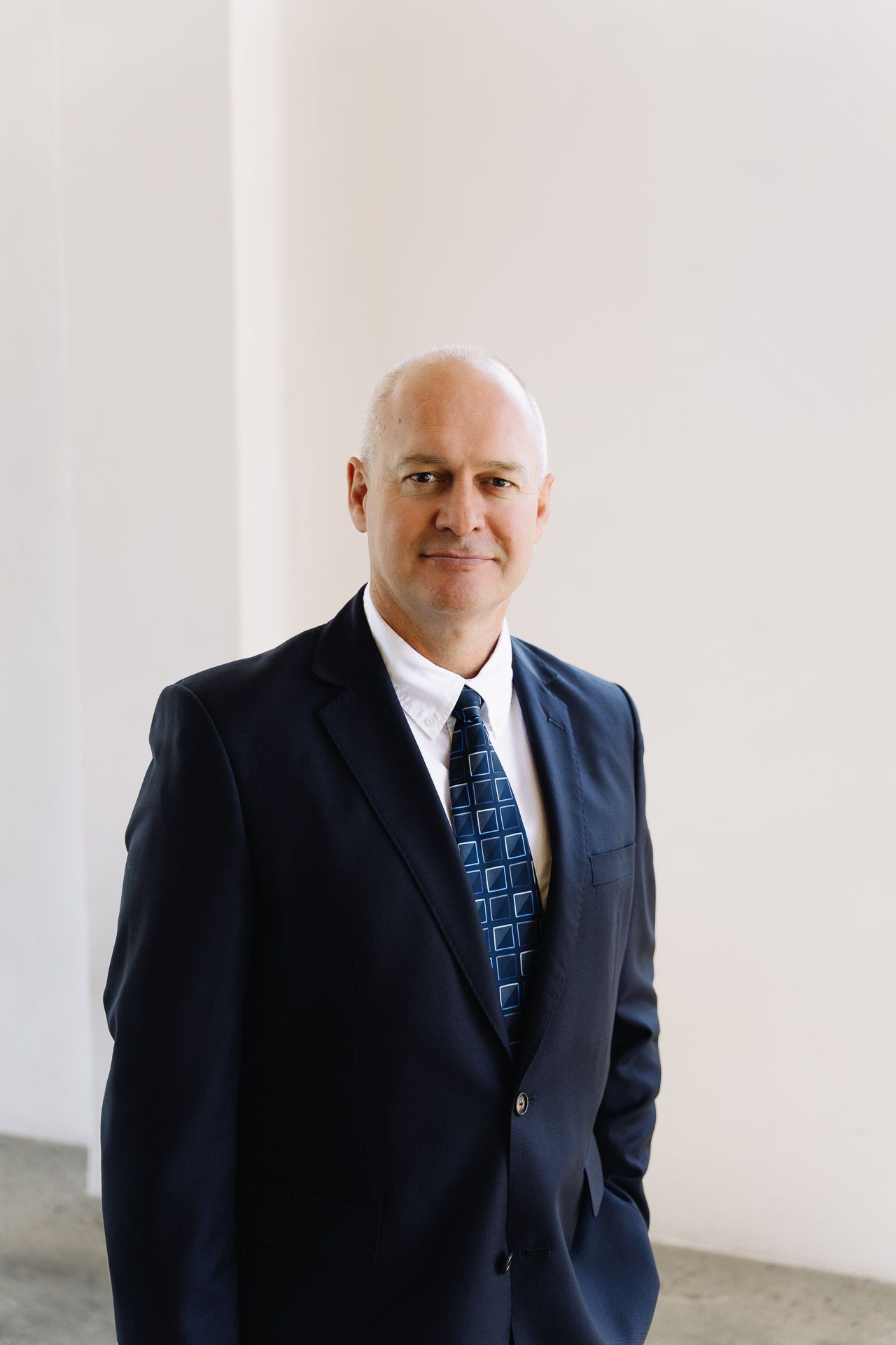 Man in a dark suit and patterned tie, smiling, standing in a white hallway.