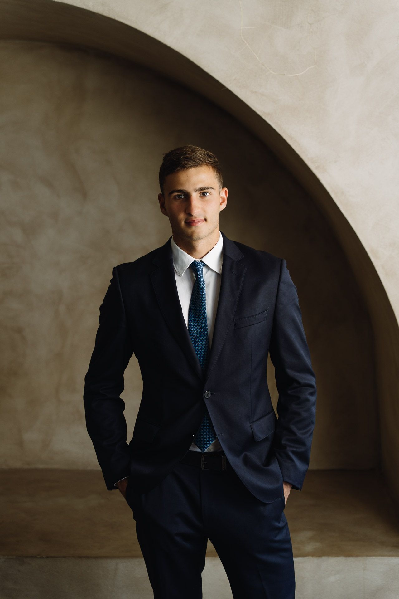 Man in navy suit, hands in pockets, stands against a curved beige wall.