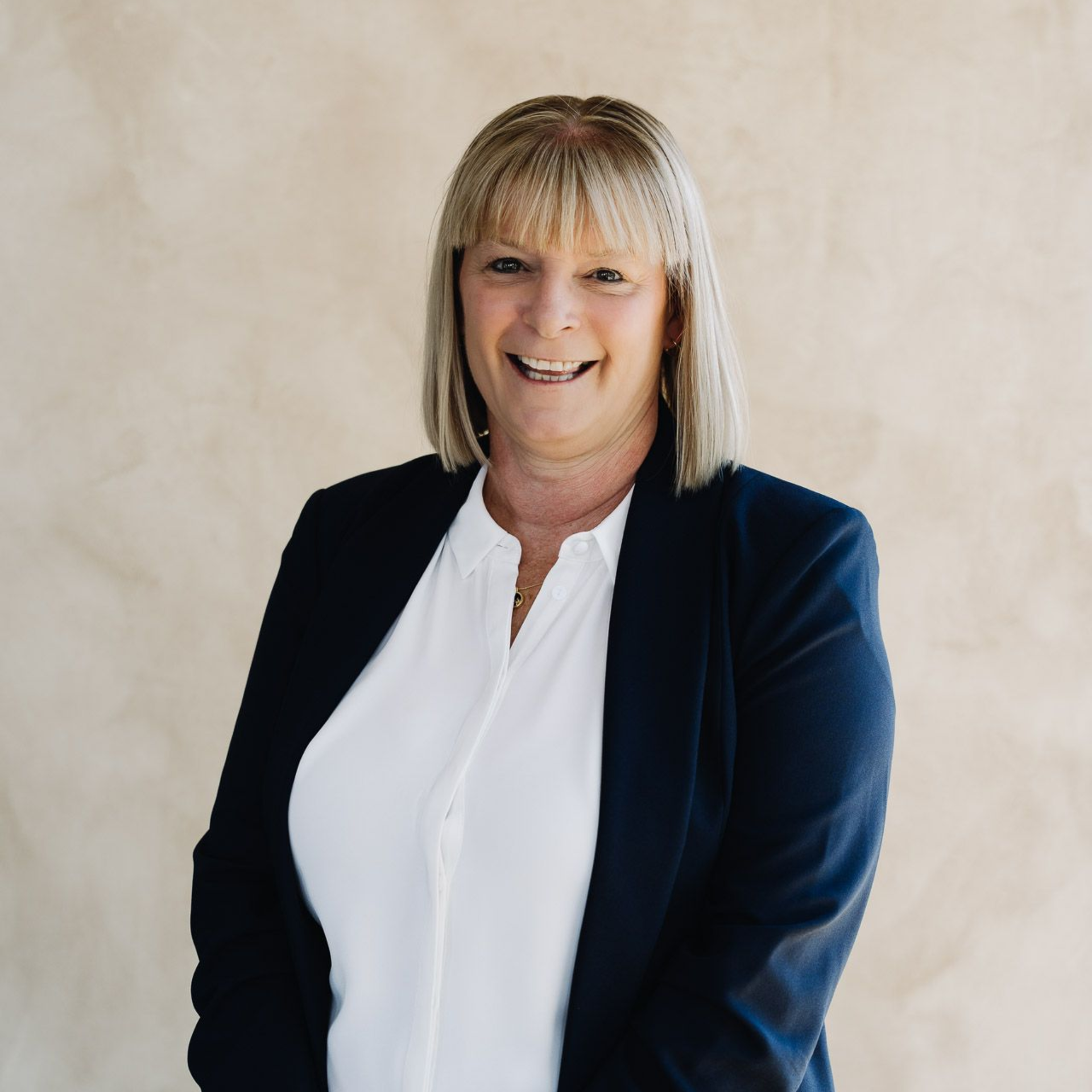 Woman in a navy blazer and white shirt smiling, against a neutral-colored wall.