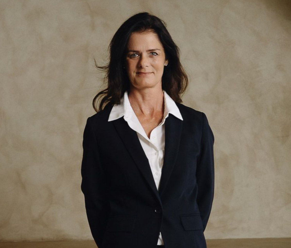 Woman in a dark blazer smiles. Pale background.