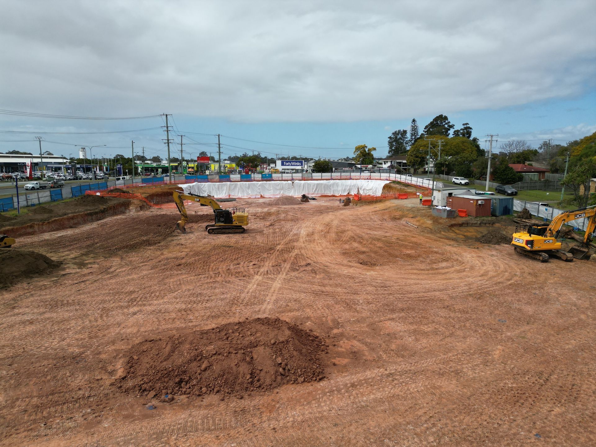 Construction site with excavators, dirt mounds, and a white fence under a cloudy sky.